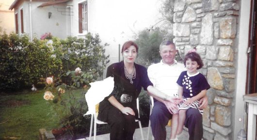Still from My Father and Qaddafi: a family group sits outdoors on garden chairs in front of a stone house with rose bushes.