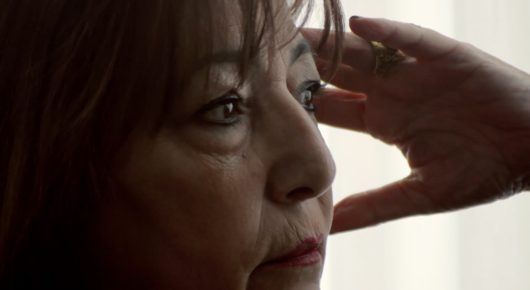 Still from My Father and Qaddafi: a close-up of an older woman with reddish-brown hair, her hand raised to shield her eyes.