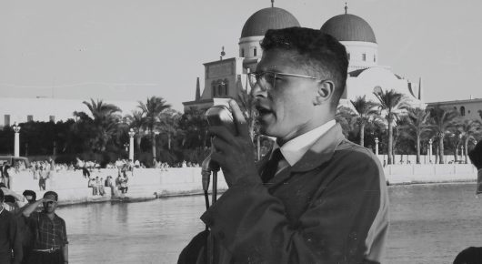 Still from My Father and Qaddafi: an archive black-and-white photograph of a man in a suit and glasses speaking at a microphone in front of a palm-lined seafront with domed buildings.