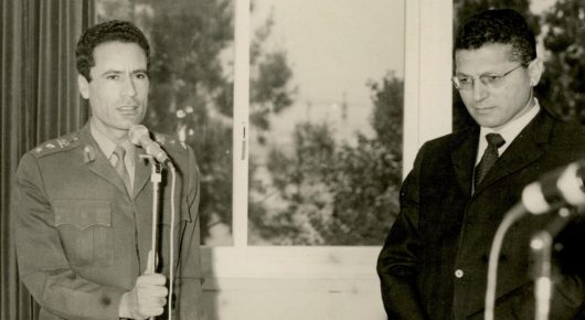 Still from My Father and Qaddafi: an archive black-and-white photograph of a man in military uniform standing at a microphone beside a man in a suit and glasses, with trees visible through the window behind them.