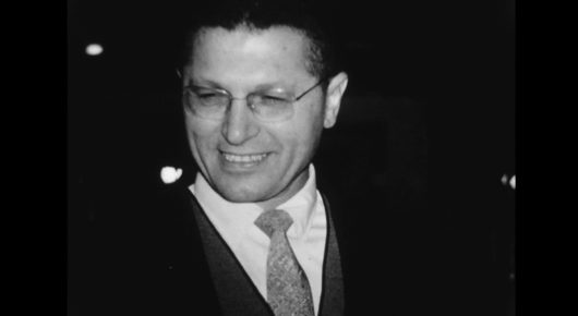 Still from My Father and Qaddafi: a black-and-white portrait of a smiling man with glasses, in a suit and tie.