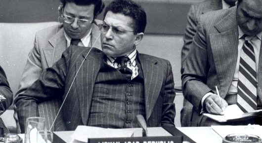 Still from My Father and Qaddafi: an archive black-and-white photograph of men in suits seated at a long table with microphones, one in glasses writing, in front of a nameplate reading Libyan Arab Republic.