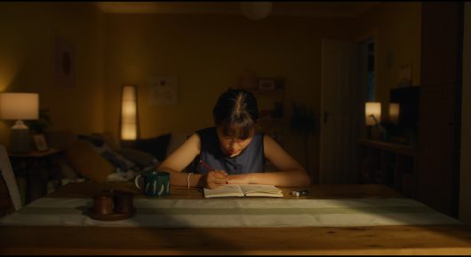 Still from Megumi: a girl in school uniform kneels at a low table in a warmly lit room, writing.