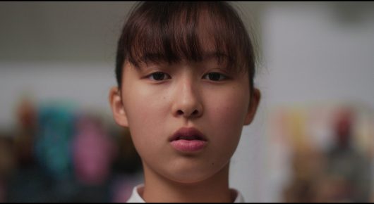Still from Megumi: a close-up of a girl with a fringe looking straight at the camera.