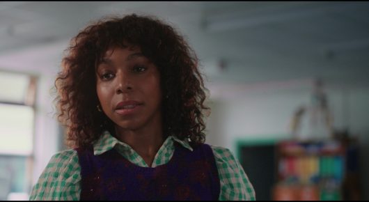 Still from Megumi: a woman with curly hair, a waistcoat and a patterned shirt looks to one side in a classroom.