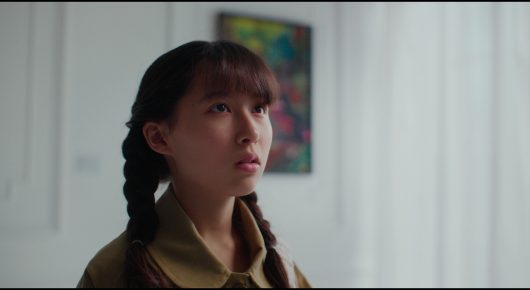 Still from Megumi: a girl with a dark plait looks up and to one side in front of a pale wall with a framed picture.