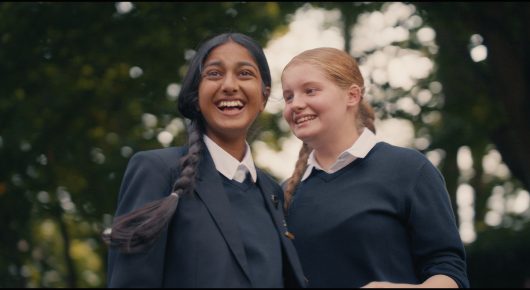 Still from Megumi: two schoolgirls in navy uniforms, one with a long dark plait, smile together outdoors.