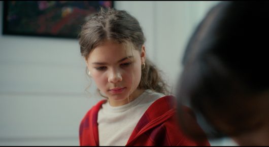 Still from Megumi: a girl with brown hair and a red cardigan looks down thoughtfully in a classroom, with another person's head at the edge of the frame.