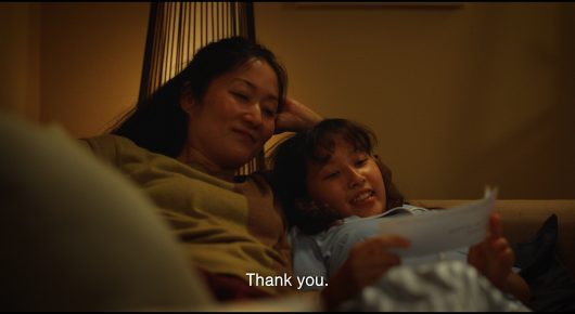 Still from Megumi: a woman with her arm round a child on a sofa, both looking at a piece of paper in warm light.