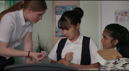 Still from Megumi: a girl in a white shirt leans in to speak to two other girls at a desk, one in a pinafore and one with dark hair and a patterned top.