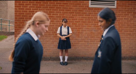 Still from Megumi: two schoolgirls in navy uniforms stand either side of a smaller girl in a pinafore, in front of a red-brick wall.