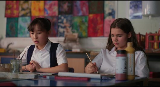Still from Megumi: two girls in school uniform sit at desks in an art room with colourful paintings on the wall behind them, one holding a paintbrush.