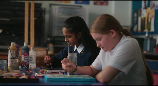 Still from Megumi: two girls sit at a classroom table painting and drawing, with art supplies in front of them.