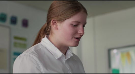 Still from Megumi: a schoolgirl in a white shirt with her hair in a ponytail looks down and speaks in a bright classroom.