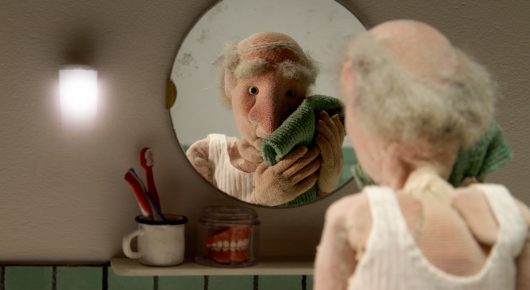 Still from Murmuration: a stop-motion puppet of an elderly man in a vest holds a cloth up to his reflection in a round mirror.