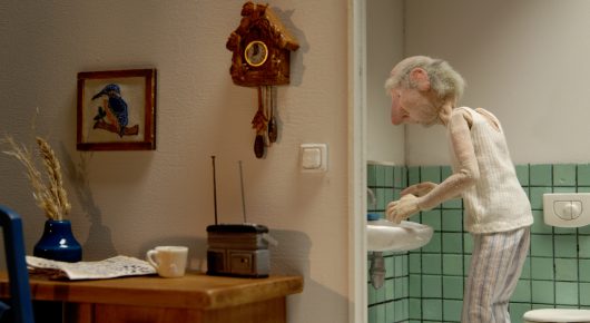 Still from Murmuration: a stop-motion puppet of an elderly person in light pyjamas washes at a sink in a tiled kitchen, with a cuckoo clock on the wall.