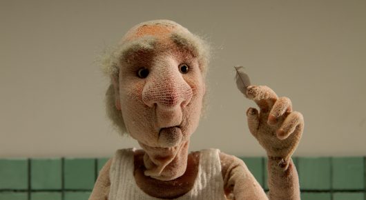 Still from Murmuration: a close-up of a stop-motion puppet of an elderly man in a vest holding up a small object.