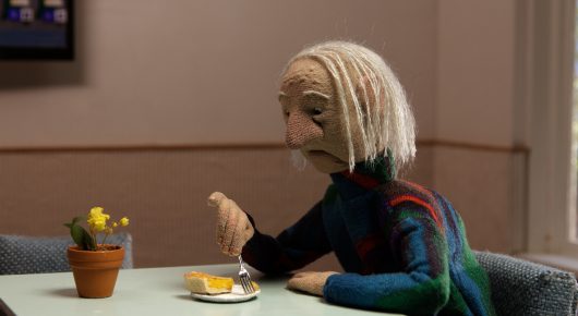 Still from Murmuration: a stop-motion puppet of an elderly person with pale hair sits at a table with a plate of food and a potted plant.