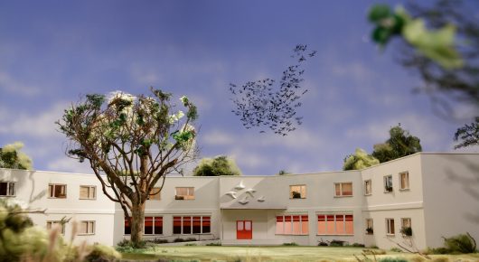 Still from Murmuration: a stop-motion model of a white building with red-shuttered windows and a large tree, with a flock of birds in the sky.
