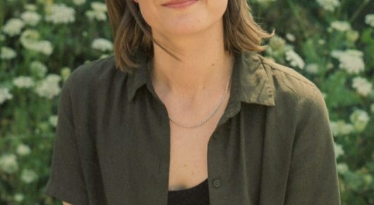 Still from Nina Roza: a woman in a green shirt and a thin necklace stands among white flowers, the frame cropped below her face.