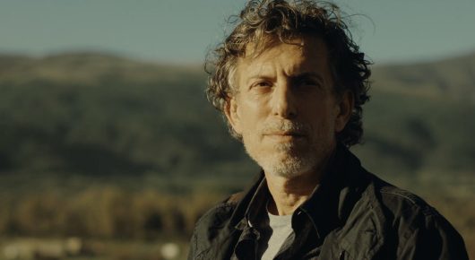 Still from Nina Roza: a man with dark curly hair and a black jacket looks at the camera, with hills behind him.