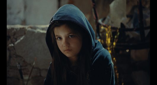 Still from Nina Roza: a girl in a dark blue hoodie looks at the camera from the shadows.