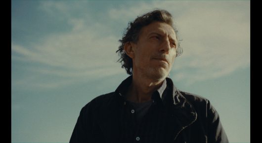 Still from Nina Roza: a man in a dark jacket stands against a pale sky, looking off to one side.