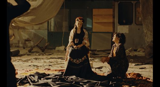 Still from Nina Roza: a woman in a long embroidered dress sits on a cloth-covered ground beside a kneeling man, in front of a wooden caravan.