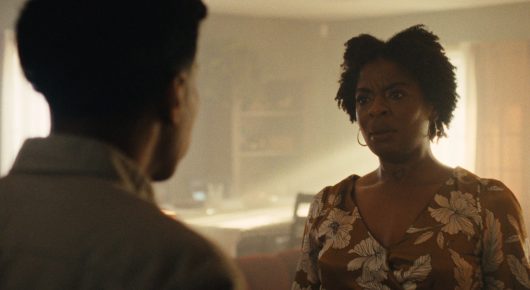 Still from One Last Night: a young man, seen from behind, faces a woman in a floral top in a warmly lit room.