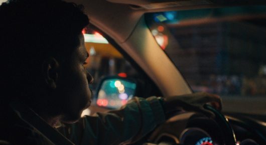 Still from One Last Night: a young man drives a car through a city at night, with coloured lights outside.