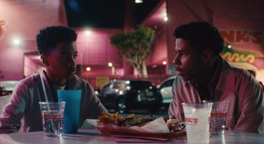 Still from One Last Night: two young men sit across a table at a fast-food restaurant with drinks and food between them, under neon lights.