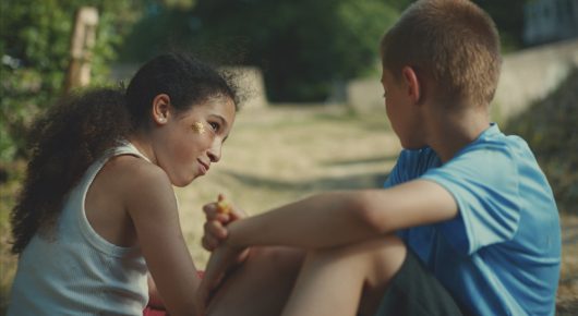 Still from Our Kingdom: a girl and a boy sit close together outdoors, the girl holding something small, with trees behind them.