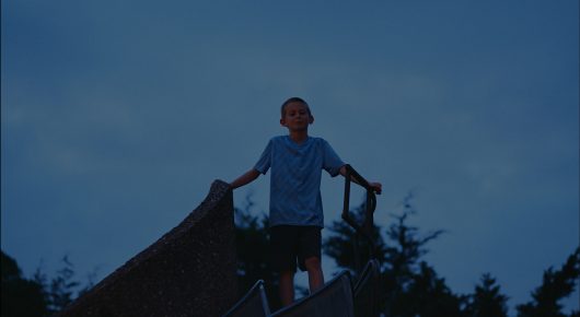 Still from Our Kingdom: a boy stands with his arms out on the edge of a roof against a deep blue evening sky.