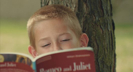 Still from Our Kingdom: a blond boy reads a copy of Romeo and Juliet, leaning against a tree.