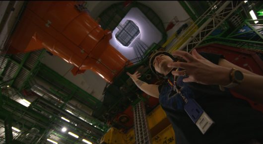 Still from The Peace Particle: a person in a dark top gestures in a machine hall with orange and white equipment, filmed from below.
