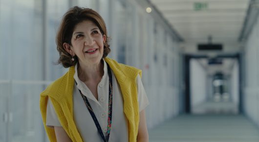 Still from The Peace Particle: a smiling woman in a yellow high-visibility vest and white shirt stands in a bright corridor.