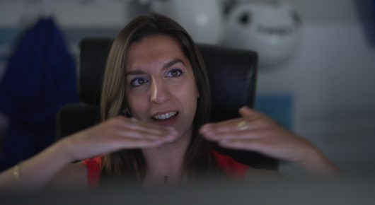 Still from The Peace Particle: a woman with long brown hair gestures with both hands as she talks, seated in a dark chair.