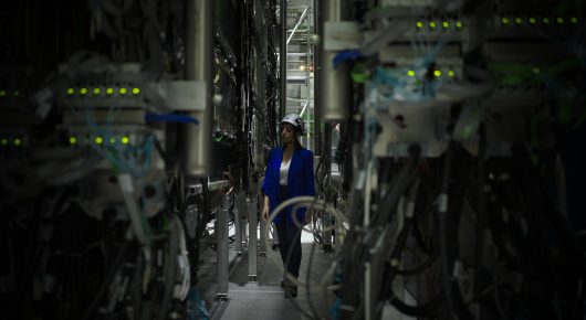 Still from The Peace Particle: a person in blue overalls and a helmet stands in a corridor of tall machinery with cables and green lights.