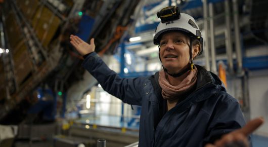 Still from The Peace Particle: a person in a white safety helmet with a head torch gestures with open arms inside a large scientific machine with blue steel supports.