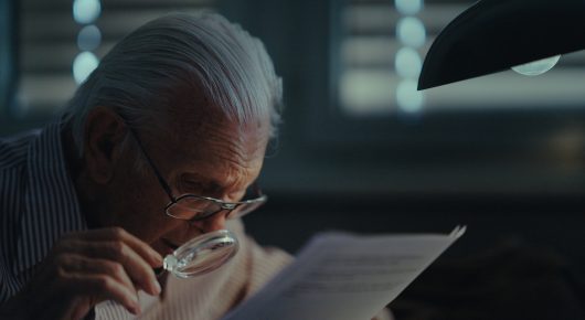 Still from The Peace Particle: an older man with white hair holds a magnifying lens over a sheet of paper at a desk lit by a lamp.