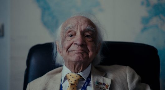 Still from The Peace Particle: an older man with white hair, a cream jacket and a yellow tie sits in a dark armchair looking at the camera, in front of a blue and white background.