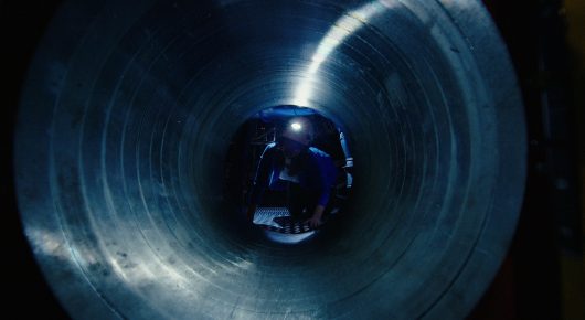 Still from The Peace Particle: a view down a large cylindrical metal tunnel lit in blue, with a small round object at its centre.