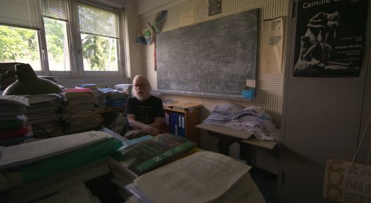Still from The Peace Particle: a cluttered office with stacks of papers and a blackboard, with a person sitting at a desk in the background.