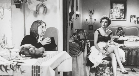 Black-and-white still from Placido: an older woman in a headscarf sits at a table in the foreground while a younger woman in a light dress sits on a bed behind her.