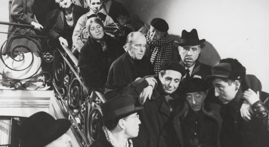 Black-and-white still from Placido: a crowd of men in hats and coats gather close together by a staircase with an ornate railing.