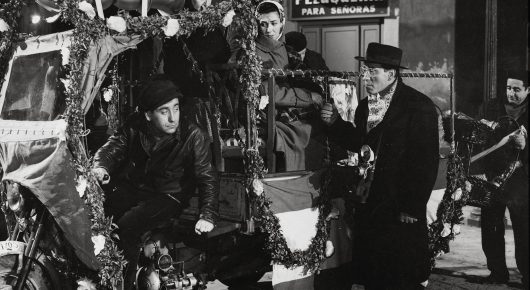 Black-and-white still from Placido: two men in coats stand by a vehicle decorated with garlands and lights on a night street, with signs behind them.