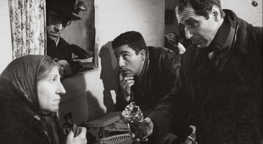 Black-and-white still from Placido: a woman in a headscarf sits close to two men in dark coats in a small room.