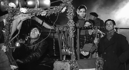 Black-and-white still from Placido: people sit and stand on a vehicle covered in lights and garlands at night.
