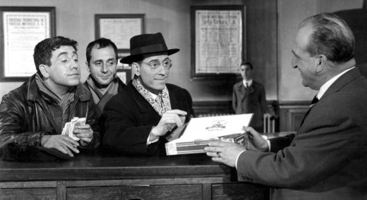 Black-and-white still from Placido: a group of men in jackets lean on an office counter, one in a hat holding a white box, as a man in a suit faces them.