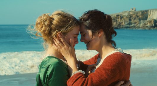 Still from Portrait of a Lady on Fire: two women in period dress, one in green and one in red, rest their foreheads together and hold each other on a beach, with cliffs behind them.